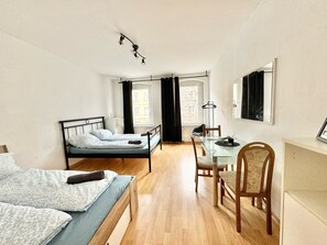 Una habitación pequeña con cama, una mesita y una silla. Hay una ventana con cortinas, una lámpara colgante y un perchero.