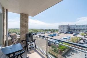 En balkon med to stole og et bord, der kigger ud over en parkeringsplads og en bygning med mange vinduer.