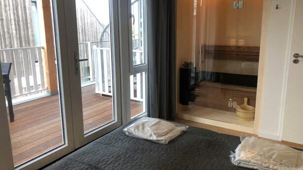 Une pièce dotée d’une grande porte-fenêtre coulissante donnant sur une terrasse en bois, d’un lit recouvert d’une couette grise et d’une salle de bain visible à travers la porte.