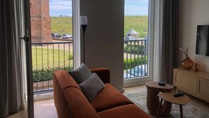 Un salon avec un canapé, une petite table ronde et un téléviseur. Il y a un balcon d’où l’on peut admirer un immeuble en briques et un espace verdoyant.
