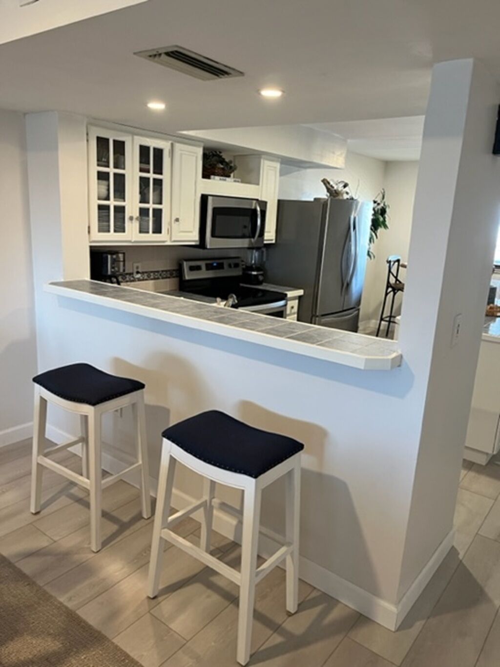 Cocina moderna con armarios blancos, microondas, estufa y refrigerador. Hay dos taburetes de bar con cojines negros.