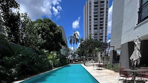 A clear blue pool with a tiled edge, surrounded by greenery and a building with a balcony. - Giardino Smart Studio 05205 · 7 km GRU Airport +Space+Pool (Guarulhos)