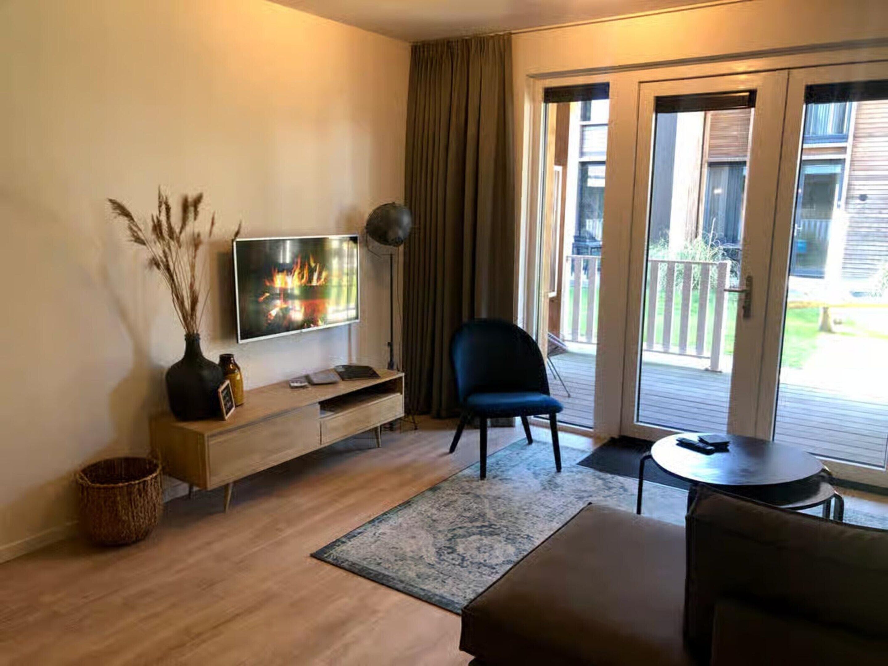Een moderne woonkamer met een flatscreen-tv, een donkerblauwe fauteuil, een klein rond tafeltje en een houten tv-meubel.