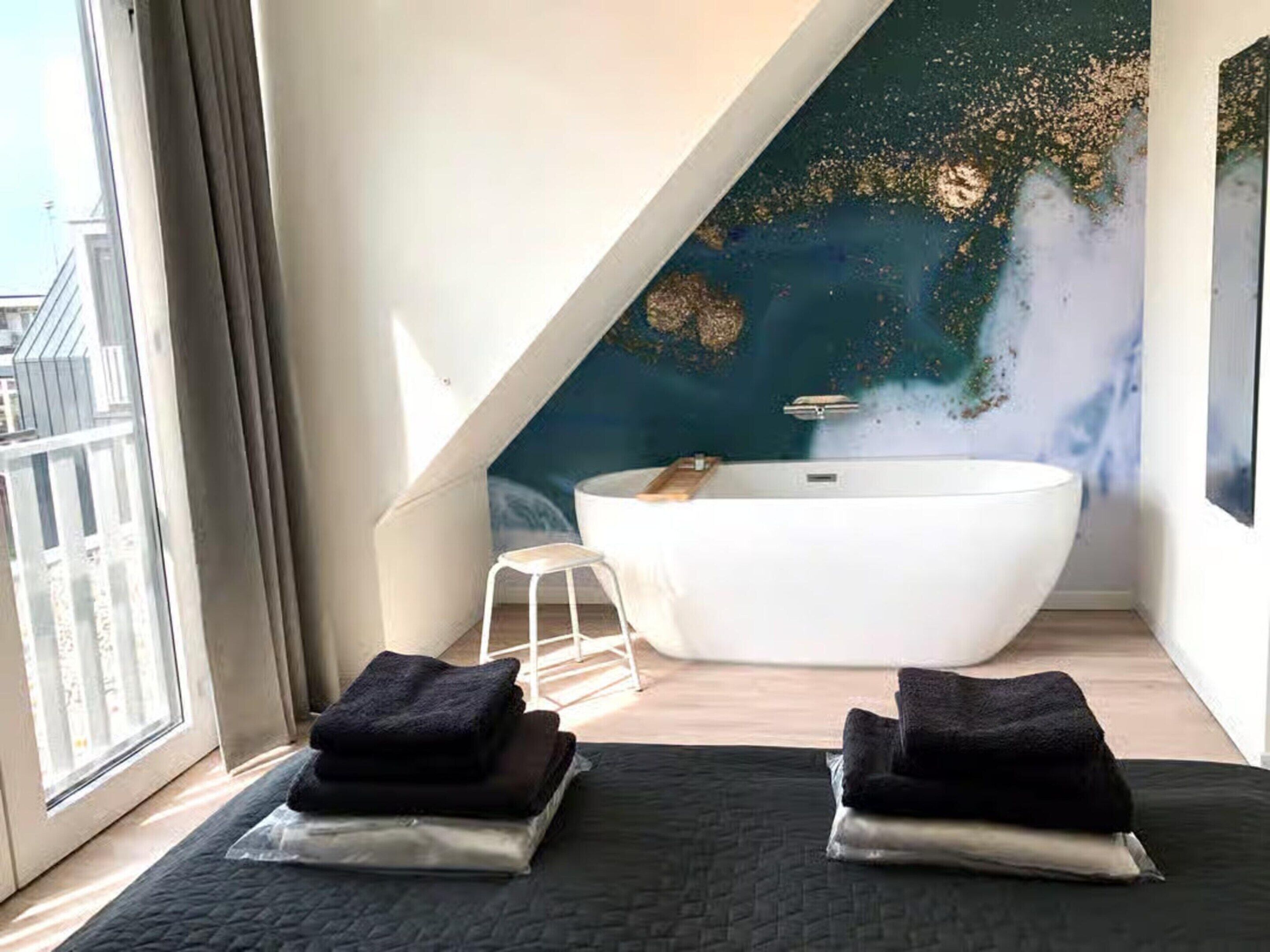 A modern bathroom with a freestanding bathtub, a small stool, and towels on a bed.