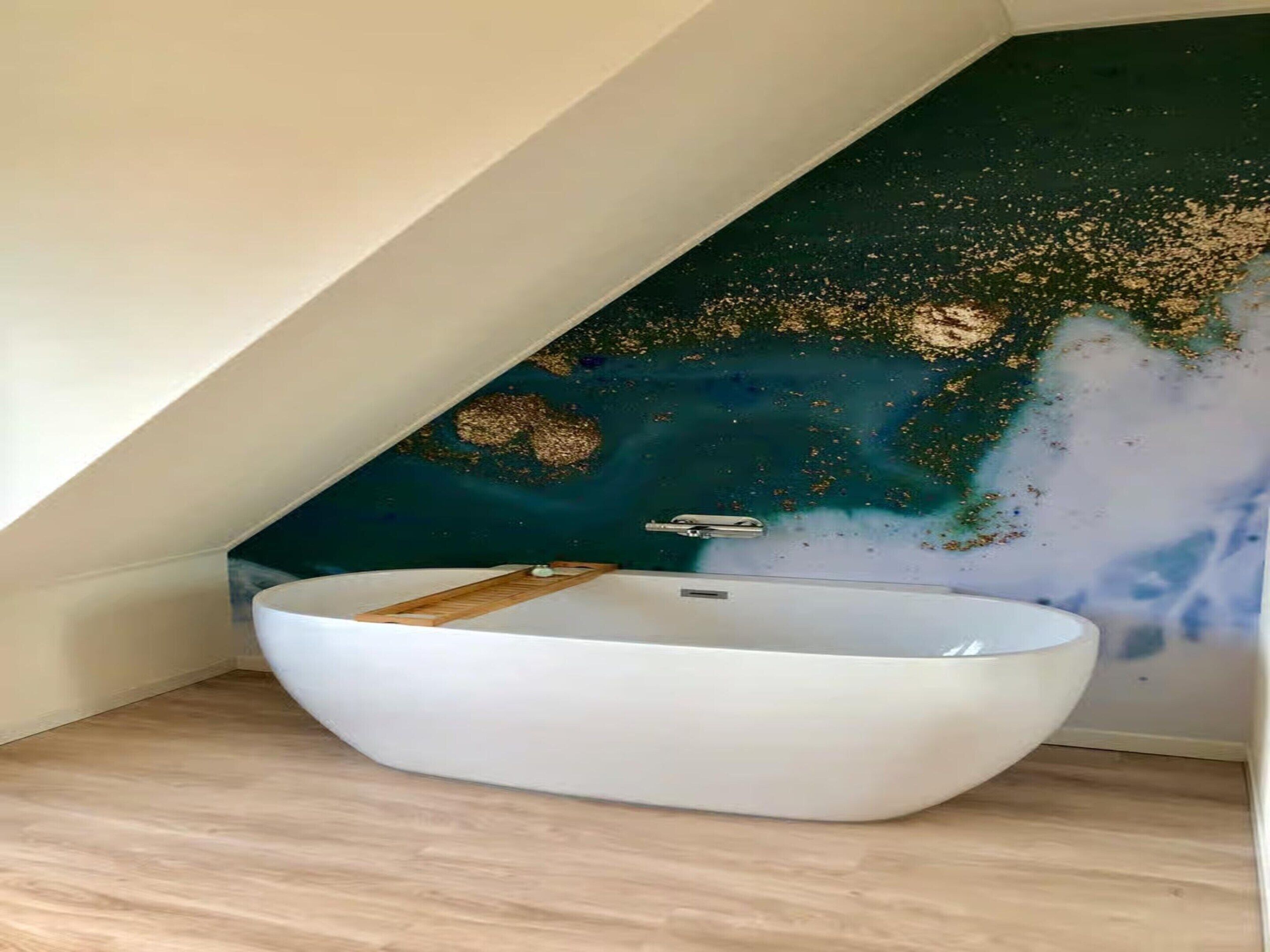 Ein modernes Badezimmer mit einer freistehenden weißen Badewanne, einer dunkel gefärbten Wand mit Goldglitzer und einem Holzboden.