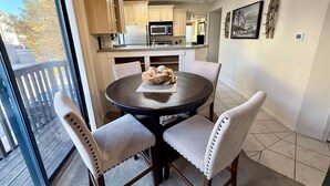 Un coin repas avec une table ronde et quatre chaises, une cuisine à l’arrière-plan et une vue à travers une porte vitrée.