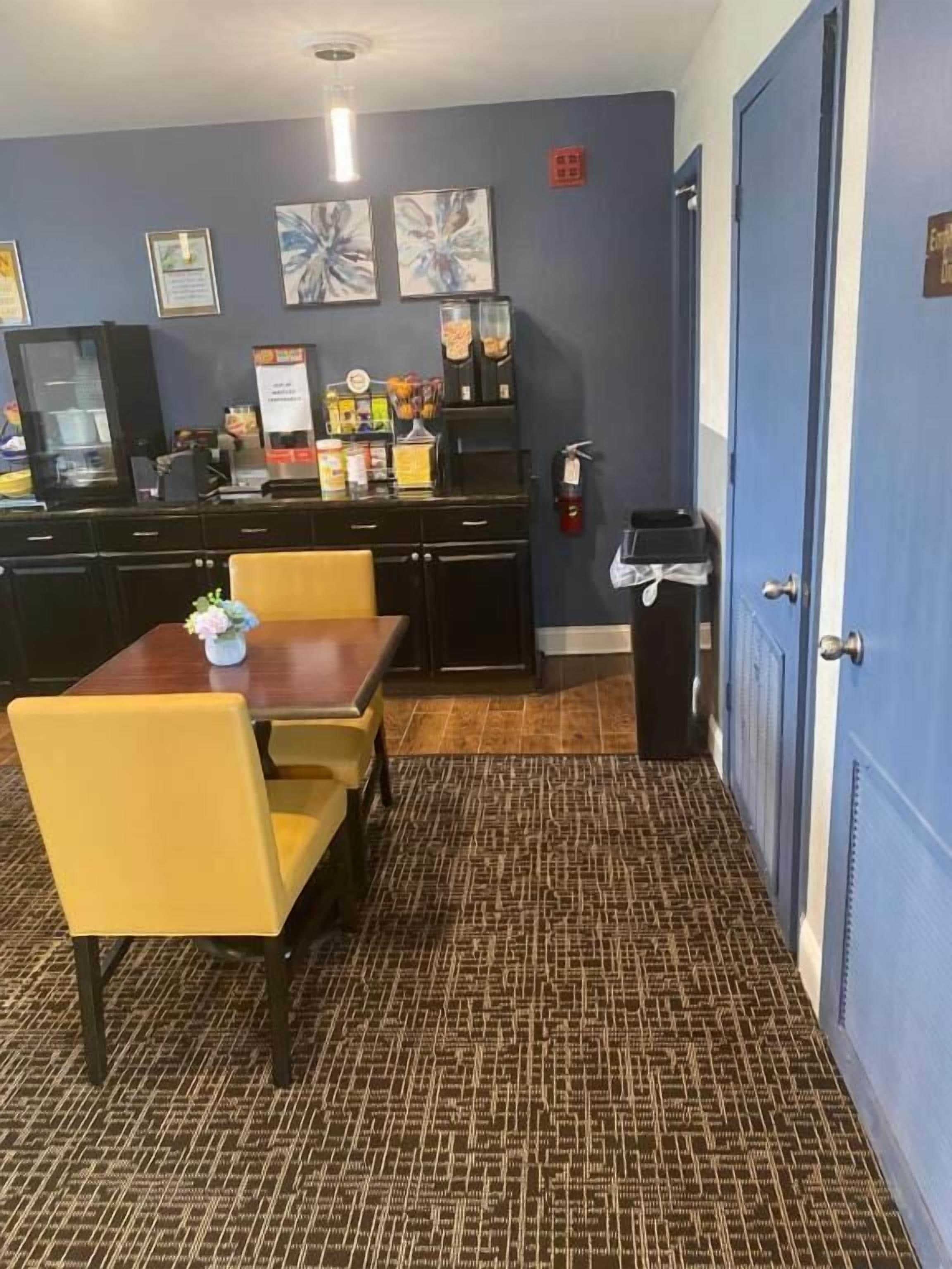 Une chambre d’hôtel compacte avec un coin repas comprenant une table en bois et des chaises jaunes, une kitchenette équipée d’un réfrigérateur et d’une machine à café, et une porte bleue.