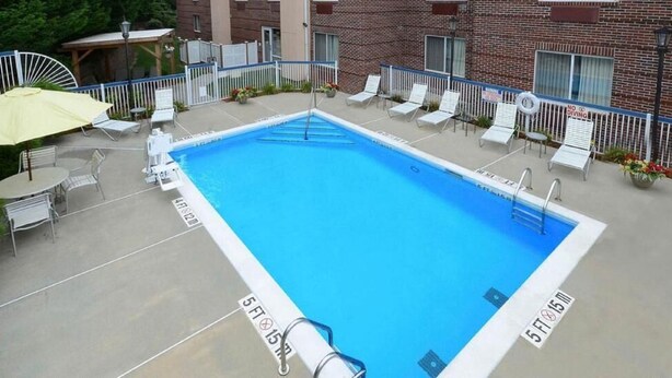 A rectangular pool with a diving board, surrounded by white lounge chairs and a yellow umbrella. - Fairview Inn Airport (Greensboro)