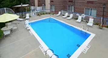 Una piscina rectangular con un trampolín, rodeada de sillas reclinables blancas y un paraguas amarillo.