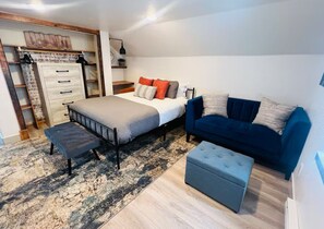 Un dormitorio con una cama, un sofá azul, un reposapiés gris, un aparador de madera y una alfombra estampada.
