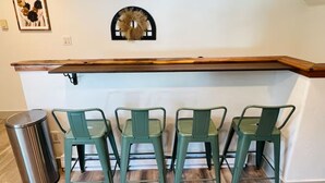 Una zona de cocina con encimera de madera, cuatro sillas verdes, un cubo de basura y una corona decorativa en la pared.