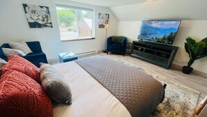 Un dormitorio con una cama grande, televisión, un sillón azul y ventana con vistas a la vegetación.