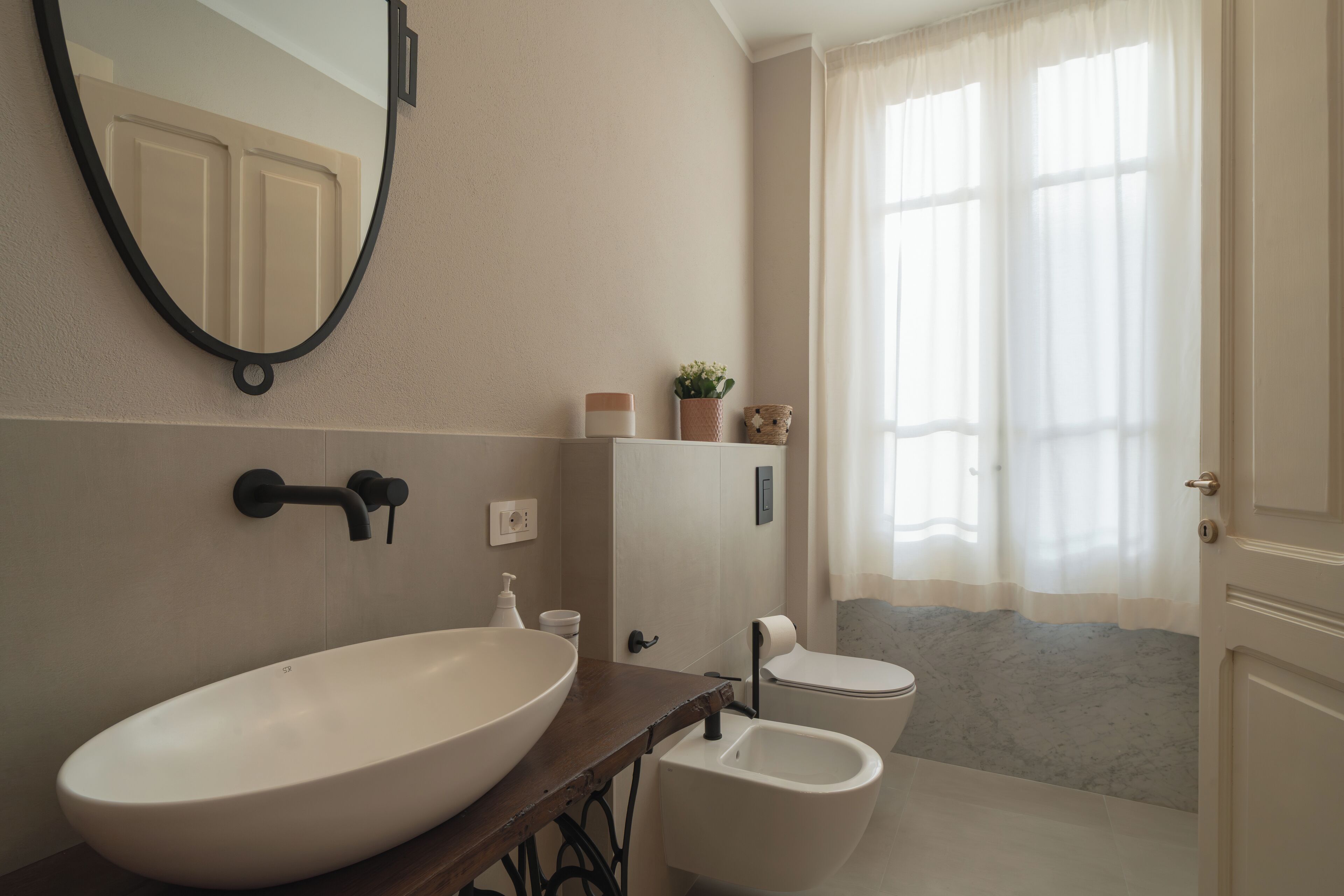 A bathroom with a large oval sink, a toilet, and a bidet. There is a mirror on the wall, a small plant, and a window with curtains.