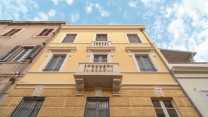 A multi-story building with a yellow facade, large windows, and a balcony with a balustrade. - Arrecramos (Cagliari)