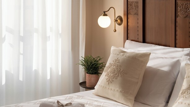 A neatly made bed with white linens and decorative pillows, a wall-mounted light fixture, and a small potted plant on a bedside table. - Arrecramos (Cagliari)