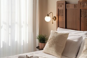 Un lit bien fait, avec du linge de lit blanc et des coussins décoratifs, un luminaire fixé au mur et une petite plante en pot sur une table de chevet.