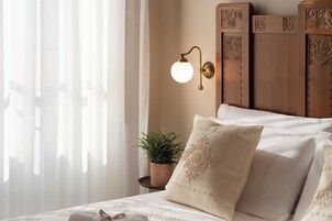 Un letto rifatto con lenzuola bianche e cuscini decorativi, una lampada a parete e un piccolo vaso con una pianta sul comodino.