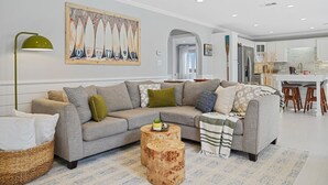 A modern living room with a grey sofa, wooden coffee table, and a painting of surfboards on the wall. - Hot Tub~Waterfront Retreat~EPIC Game Room~Private (Beaufort)