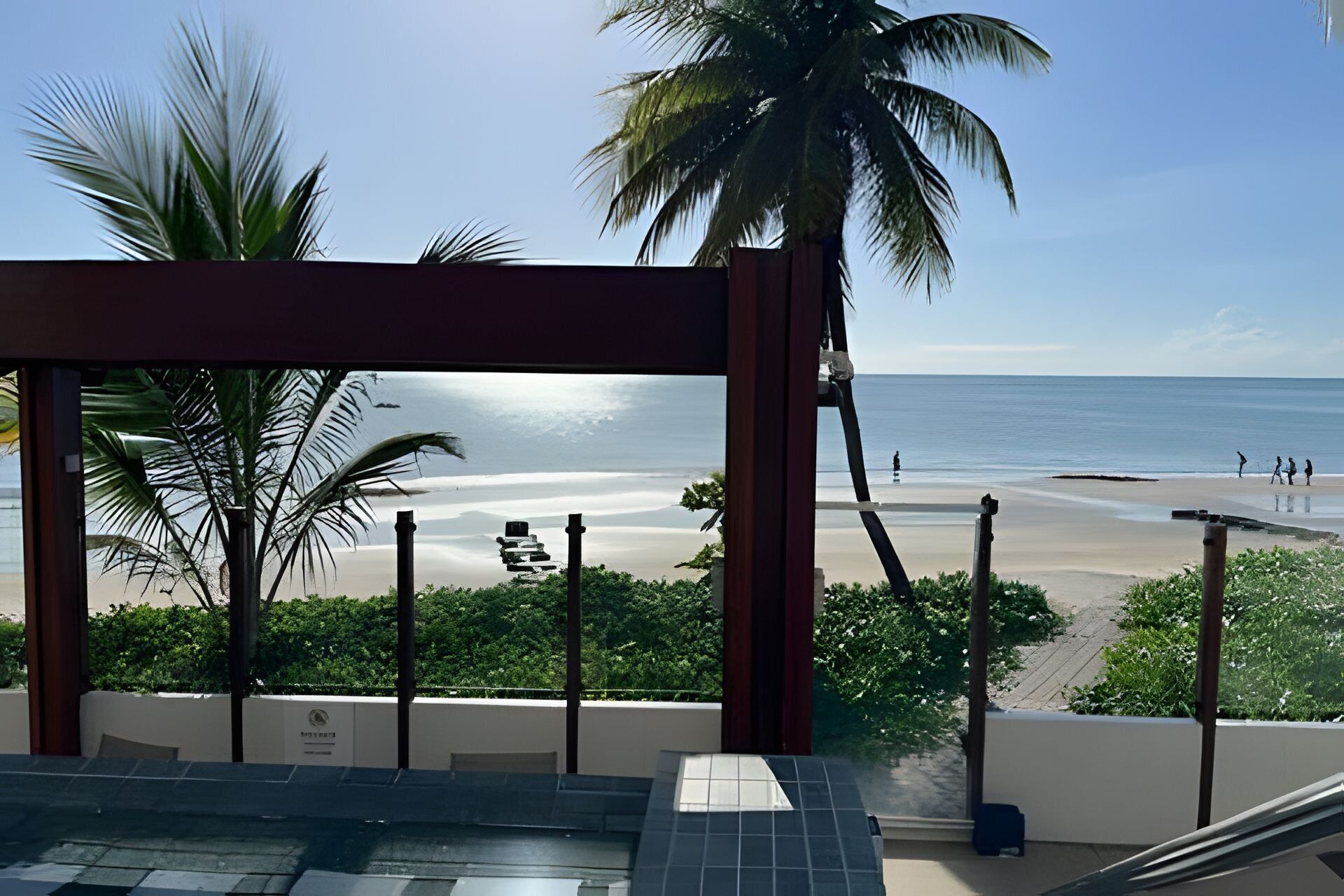 Vista de uma praia a partir de um espaço coberto, com palmeiras e céu claro.