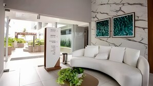 A modern interior with a white curved sofa, a marble wall, and two abstract paintings. - Flat pé na areia 406 Branco Haus  (João Pessoa)