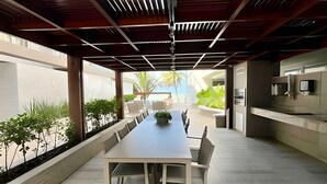 A modern outdoor dining area with a long table and chairs, a built-in grill, and a view of the outdoors. - Flat pé na areia 406 Branco Haus  (João Pessoa)