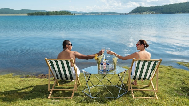 Dos personas brindando junto a una mesa de vidrio con vista a un lago, orillas cubiertas de hierba y colinas al fondo.