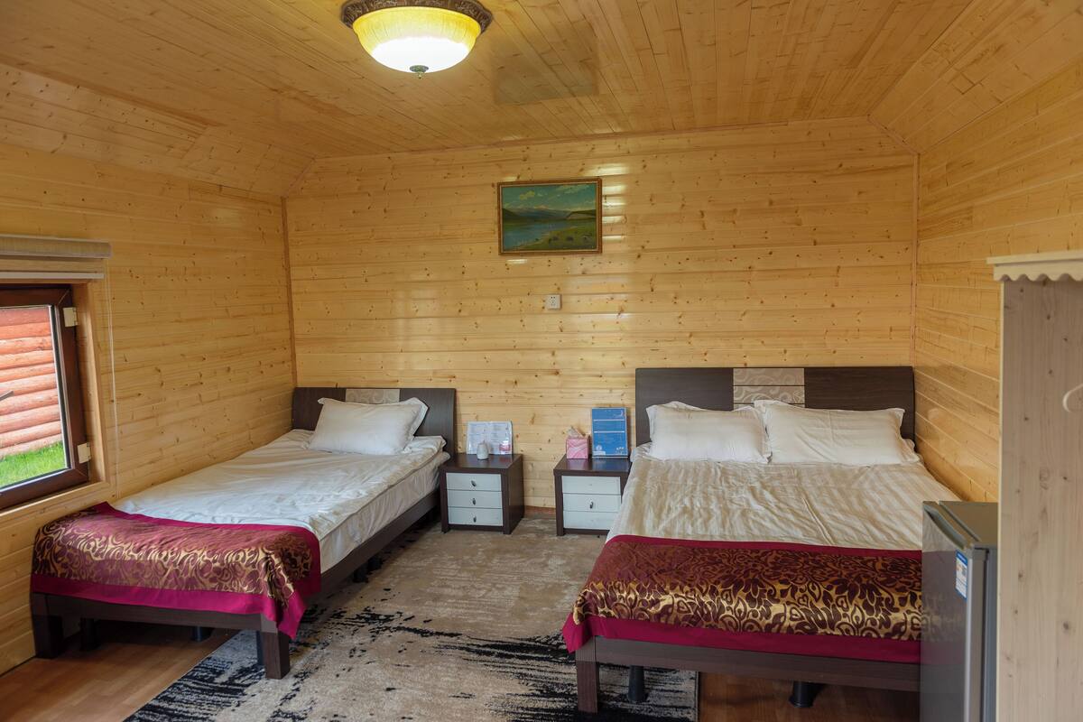 A wooden cabin bedroom with two beds, a nightstand, a refrigerator, and a painting on the wall.