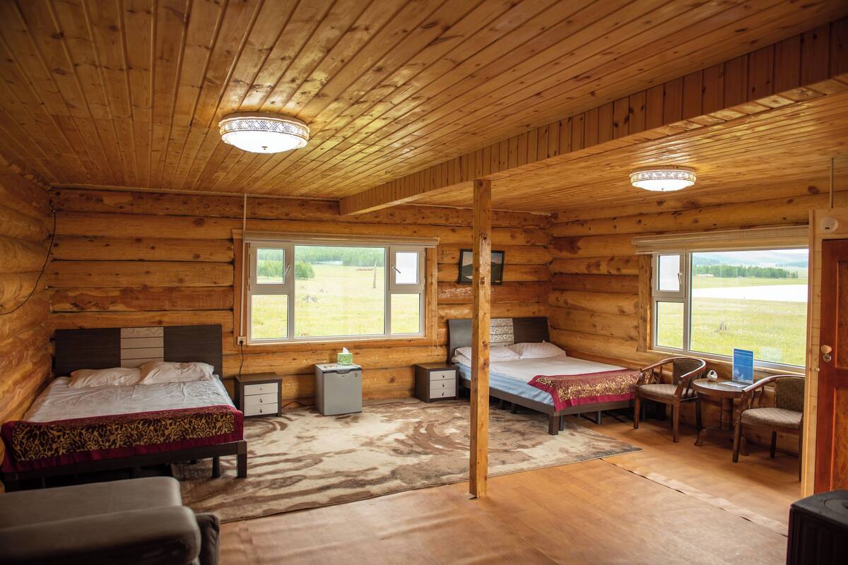 A log cabin bedroom with two beds, a desk, chairs, and a view of the outdoors.