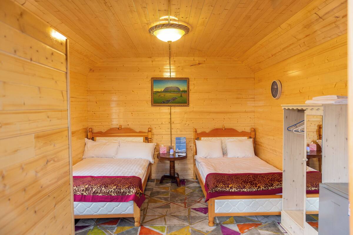 A wooden cabin room with two beds, a small table, a clock, and a painting on the wall.