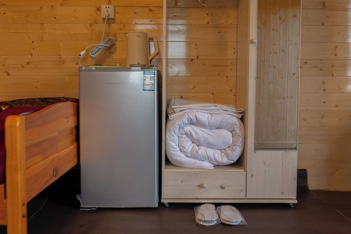 A compact room with a bed, a small refrigerator, a heater, and a storage cabinet.