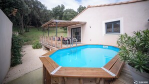 En swimmingpool med træterrasse, en overdækket terrasse og udsigt til træer samt en bygning i baggrunden.