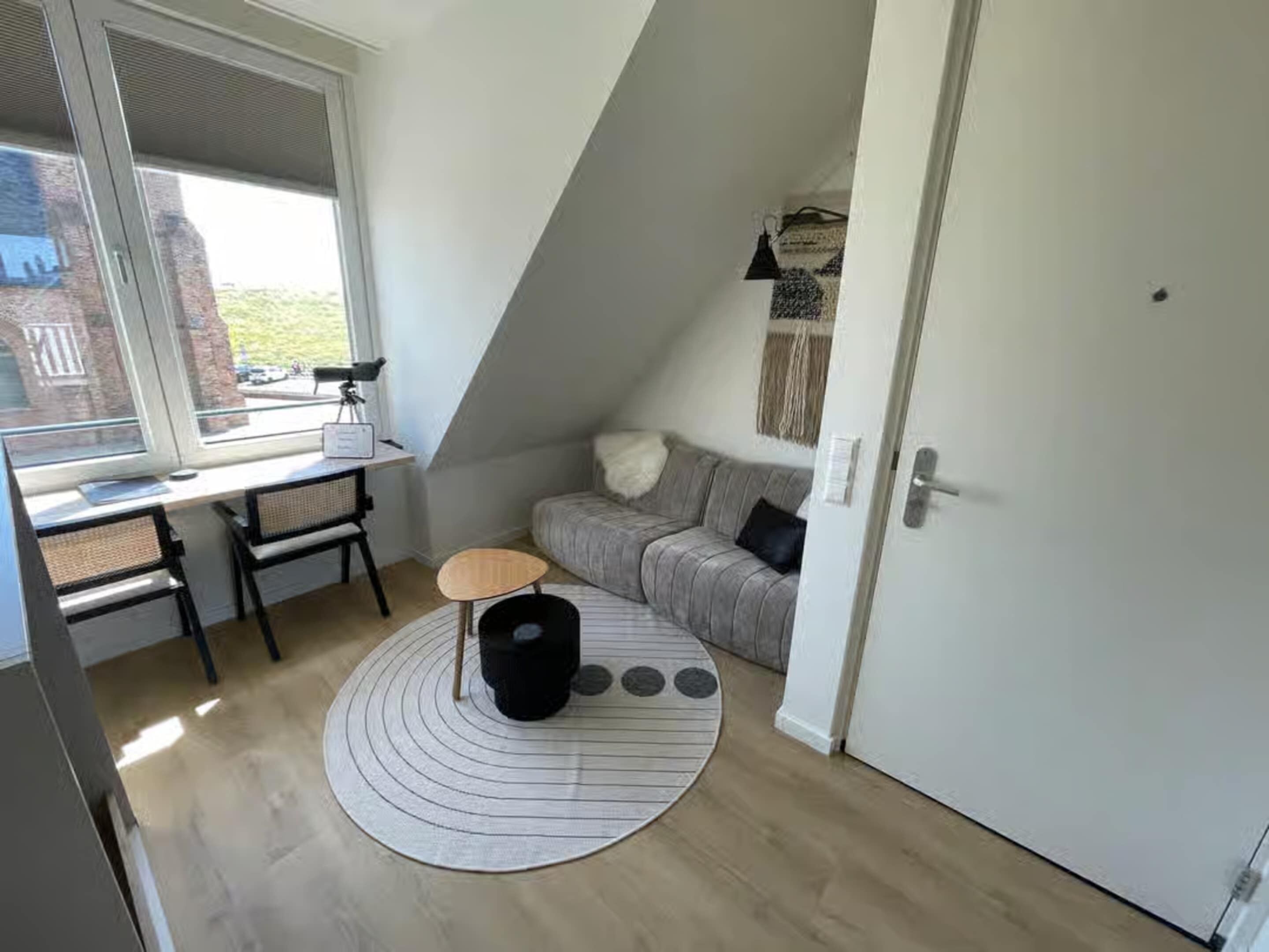 A small, cozy room with a sloped ceiling, a desk with a chair, a sofa, and a circular rug.