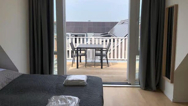 A hotel room with a bed, a table, and a view of a balcony with a railing and a building in the background. - Lodge With a Magnificent View of the Dunes (Zoutelande)