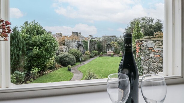 Vista desde una ventana a un jardín cuidado con un muro de piedra, un sendero y un edificio con arcos. Dos copas de vino y una botella están sobre una mesa en primer plano.