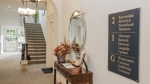 Zona de recepción de hotel con una escalera, un espejo, un escritorio de madera con un florero y un cartel con los números y nombres de las habitaciones.