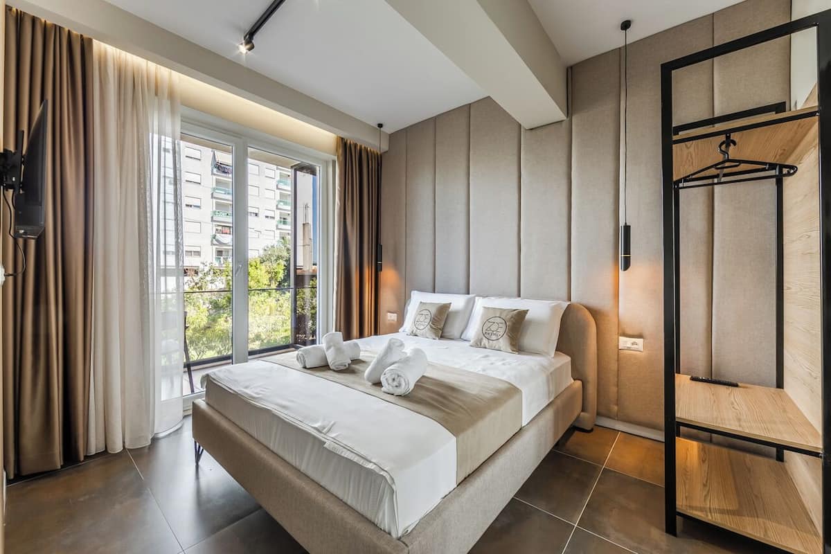 A modern hotel room with a large bed, a view of the city, and a wooden shelf.