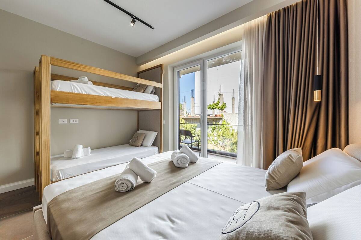 A modern hotel room with a bunk bed, a large bed with a headboard, and a balcony with curtains.