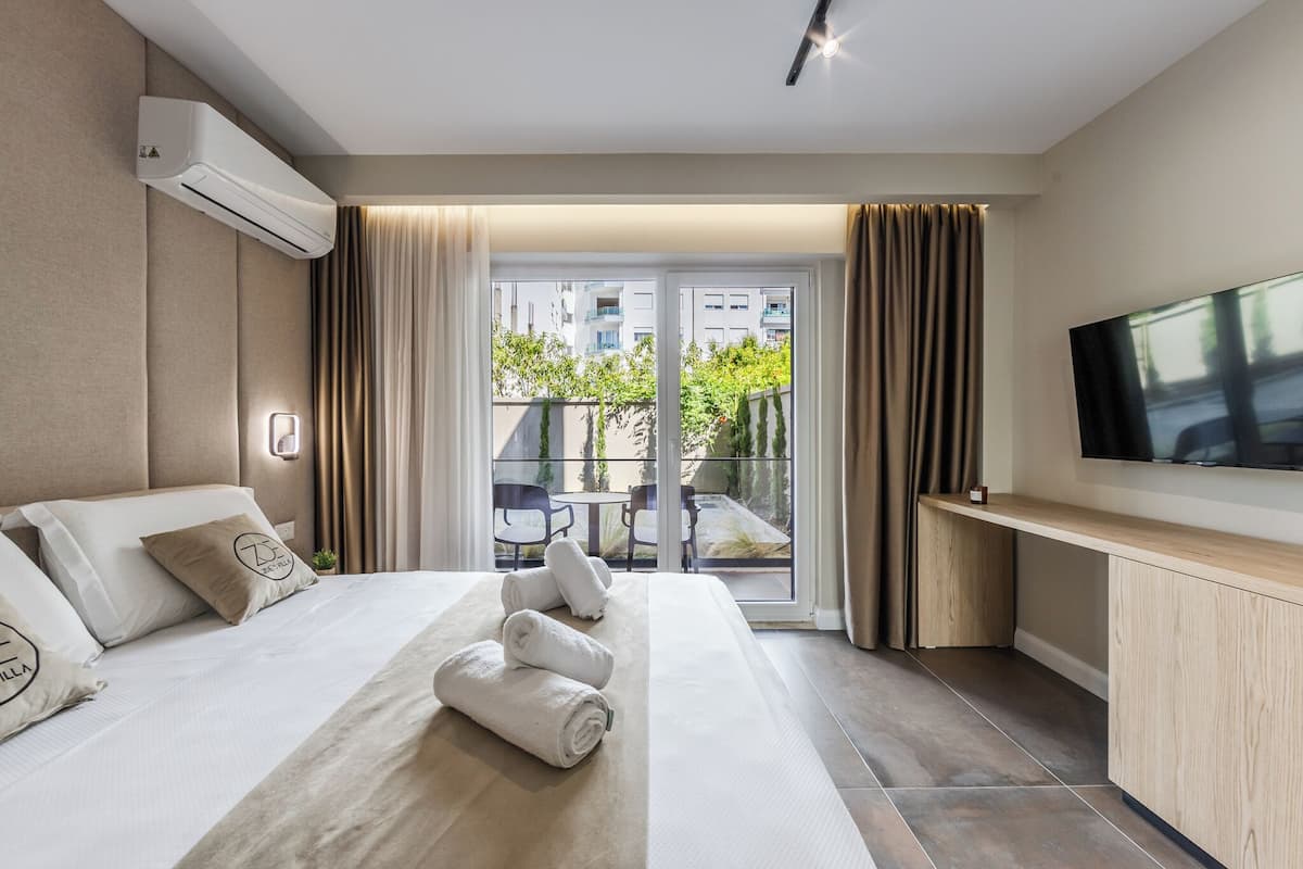 A hotel room with a bed, a TV, a balcony with a view of buildings and greenery, and a modern design.