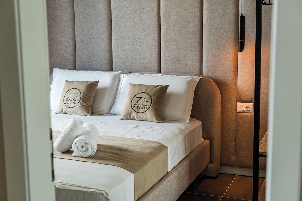 Cama bem arrumada com duas almofadas, uma toalha dobrada e luminária fixada na parede.