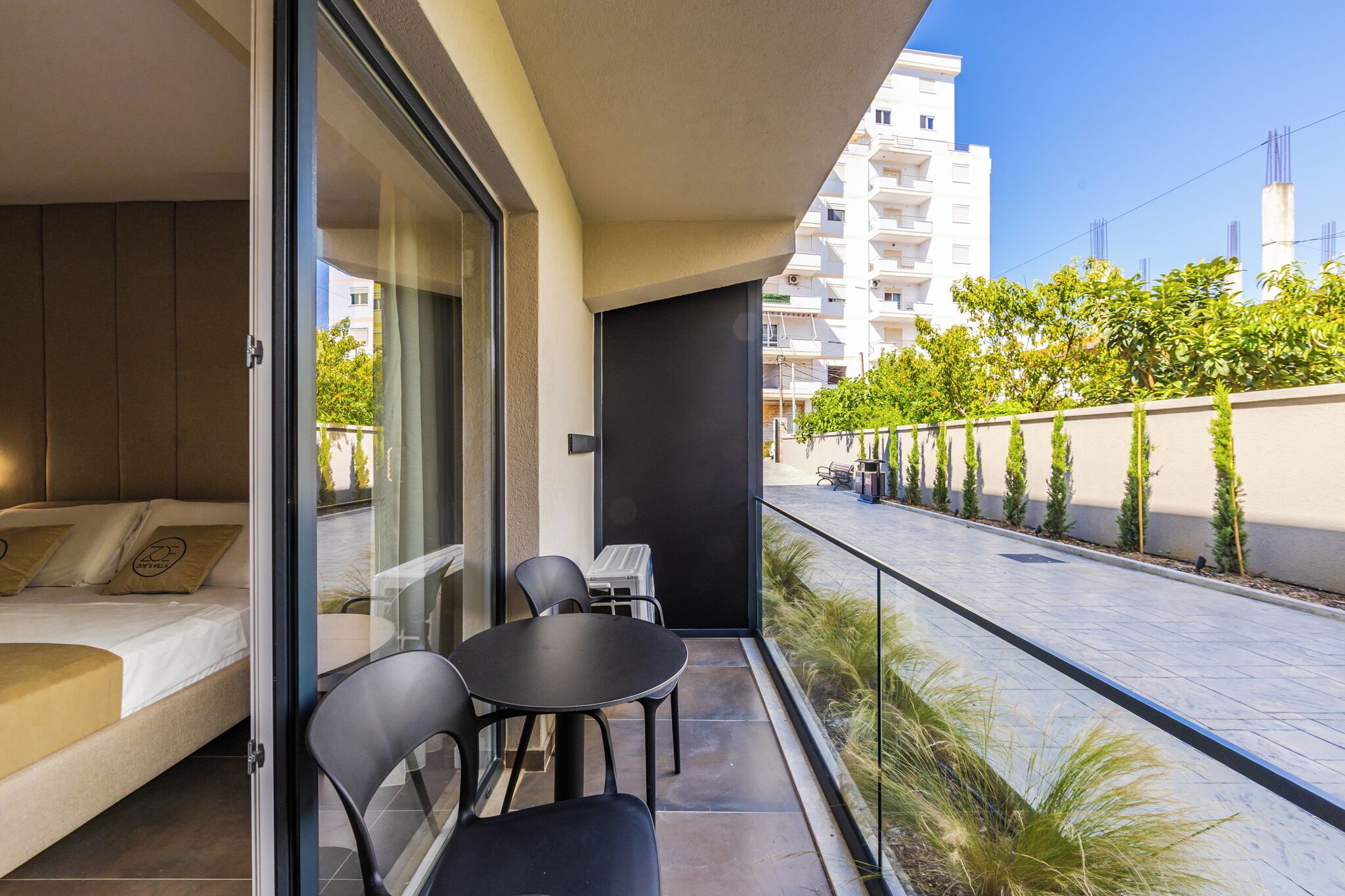 A balcony with a bed, a table, and chairs. There is a view of a street with trees and a building in the background.