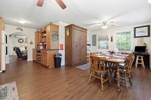 A spacious living area with a dining table, wooden chairs, a kitchen with wooden cabinets, a ceiling fan, and a window with blinds. - Goldilocks! Whittier Cabin w/ Private Hot Tub! (Whittier)
