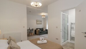 A modern hotel room with a large bed, a sitting area with a sofa and table, and a glass door leading to a shower area. - Derwent (Ulverston)