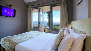 A hotel room with a bed, a TV mounted on the wall, and a view of the outdoors through a large window. - Mountain Club Resort (Chinnakanal)