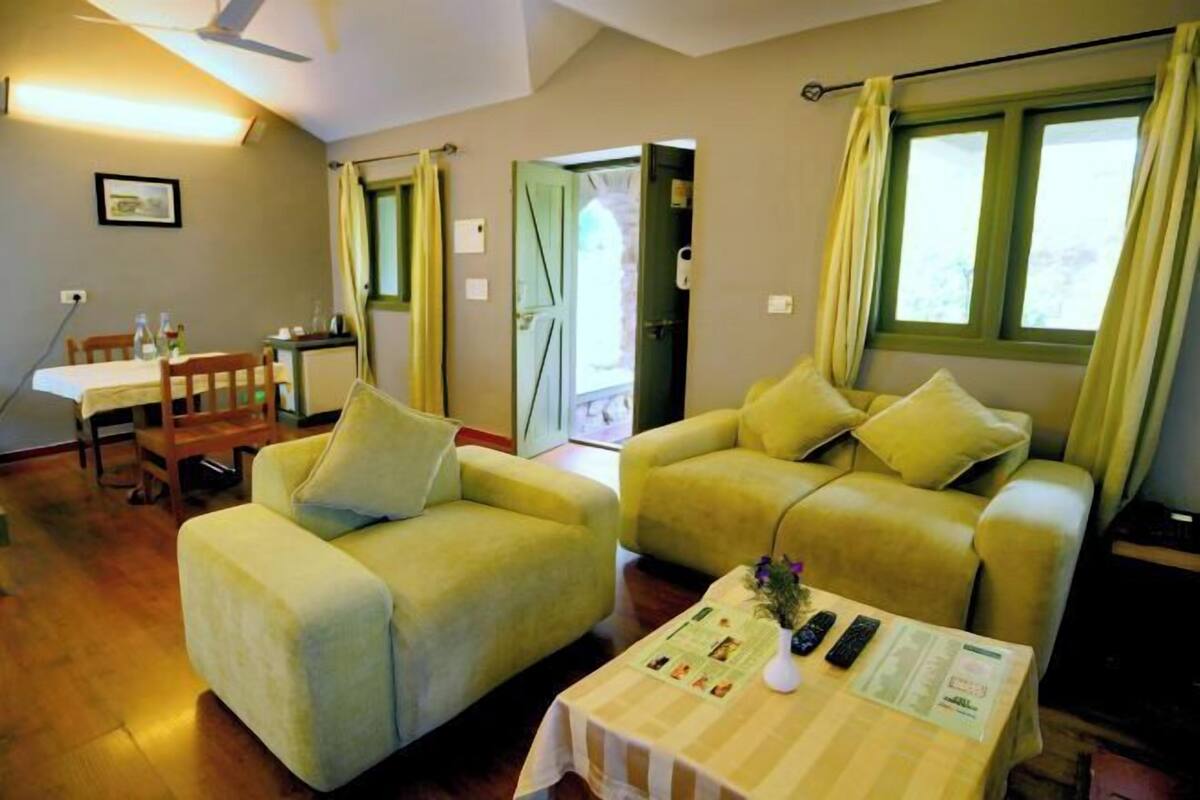 Una habitación con dos sofás amarillos, una mesa de comedor con sillas, una mesita con un florero y controles remotos, y una ventana con cortinas.