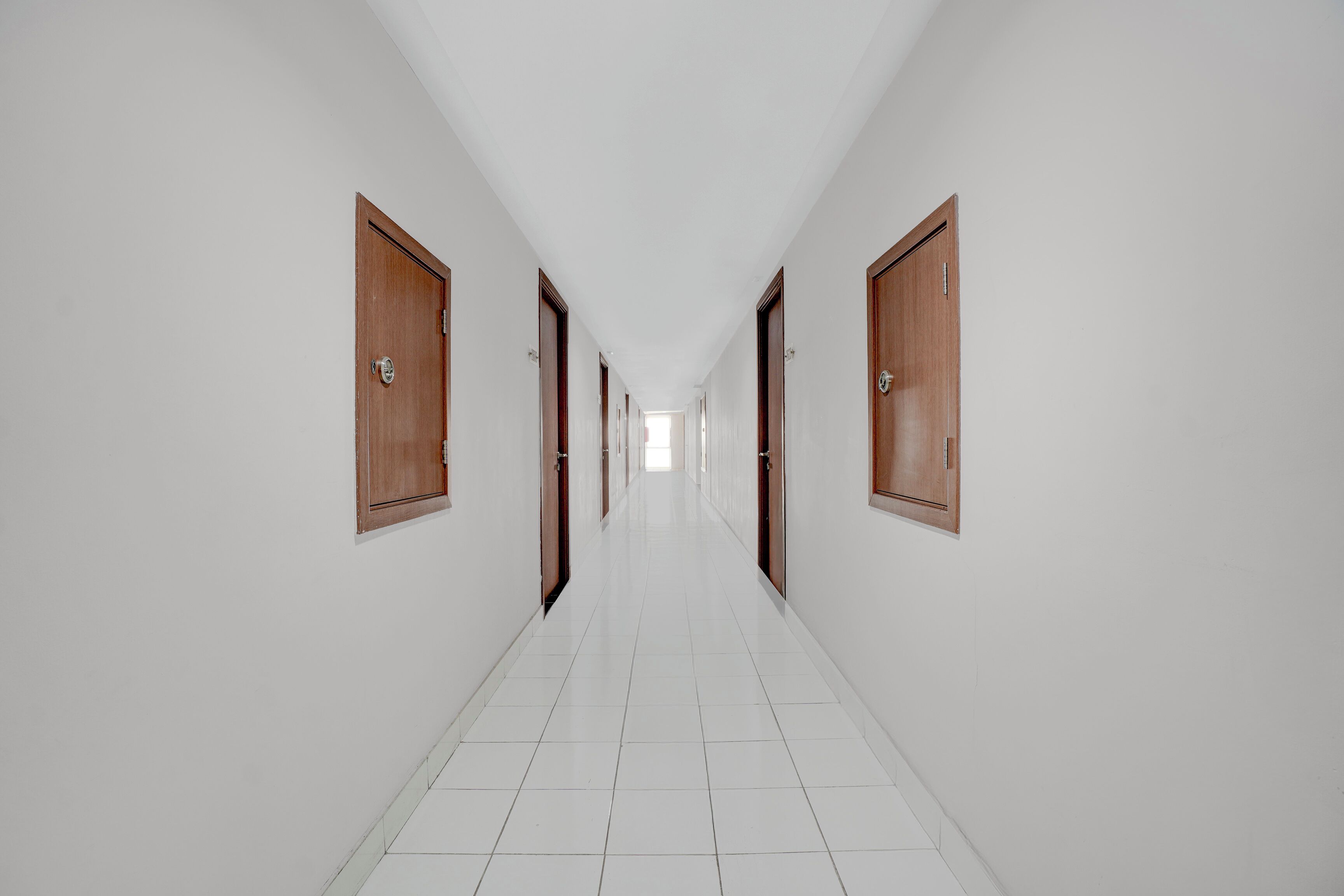 Un long couloir étroit, avec un sol carrelé de carreaux blancs et des portes en bois de chaque côté.