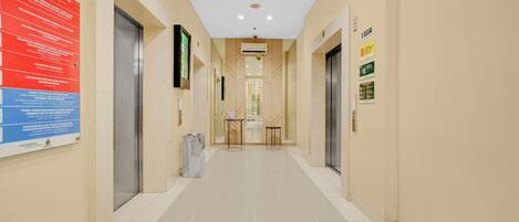 A clean, well-lit hallway with two elevators, a trash bin, and informational signs on the walls.