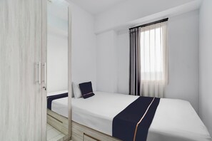 Un lit simple avec un matelas blanc, une couvre-lit bleu marine et une tête de lit en bois. Une fenêtre ornée de rideaux légers laisse entrer la lumière naturelle dans la pièce.