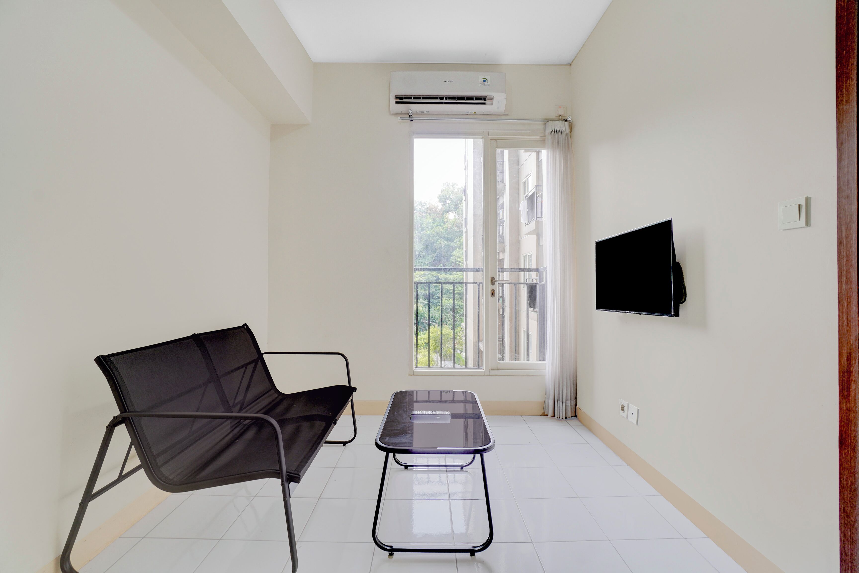 A room with a black chair, a small table, a TV mounted on the wall, and a window with a view of greenery.