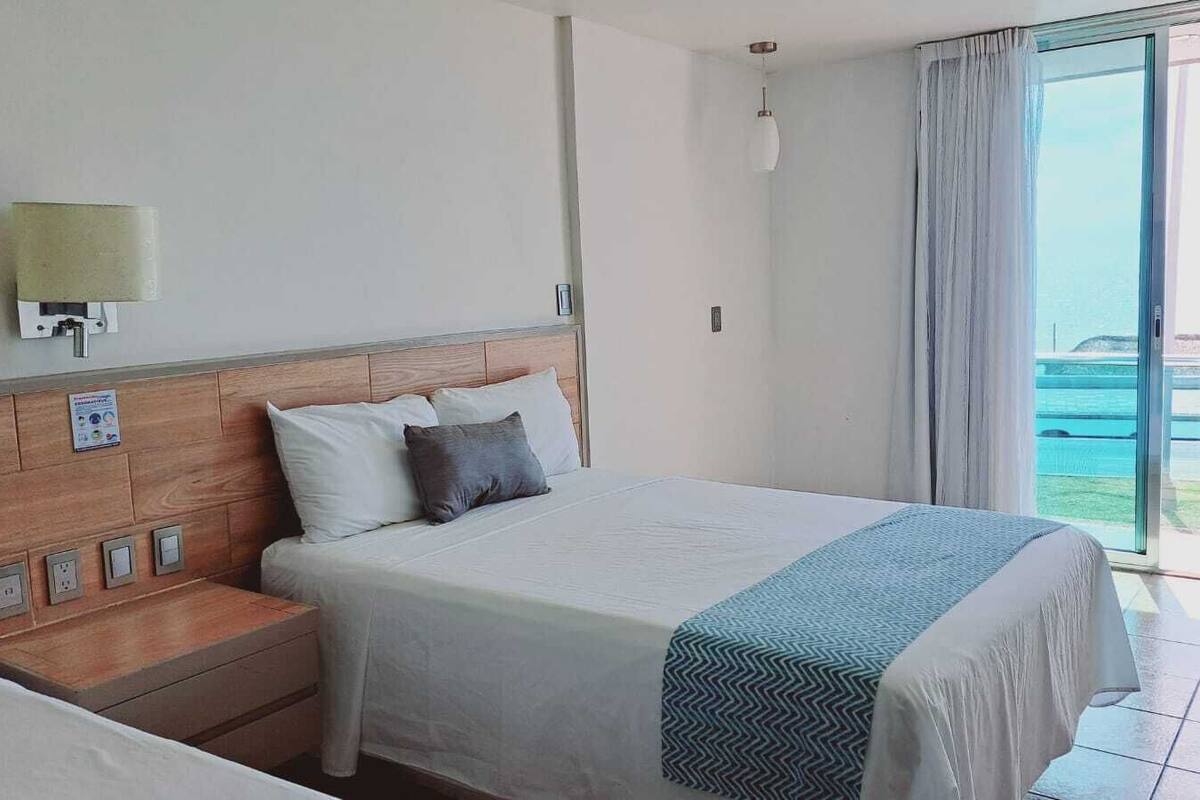 Una camera d'hotel con un letto, un comodino, una lampada a muro e vista sul mare attraverso un'ampia finestra.