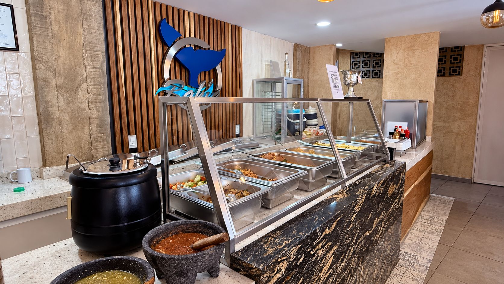 A buffet area with various food containers, a coffee station, and a decorative wall with a whale logo.